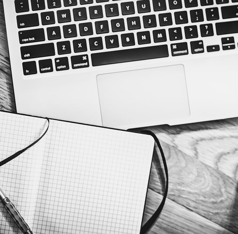 Black and white flat lay of a laptop, graph paper notebook, pen, and coffee on a wooden desk.