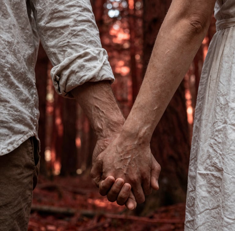 Intimate close-up of a couple holding hands in a deep redwood forest, shafts of dramatic sunlight piercing through the canopy, highlighting creased linen and rich skin tones.