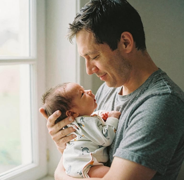 A tired but smiling father holding his newborn baby close to his chest under soft morning window light, warm domestic atmosphere, 35mm film look