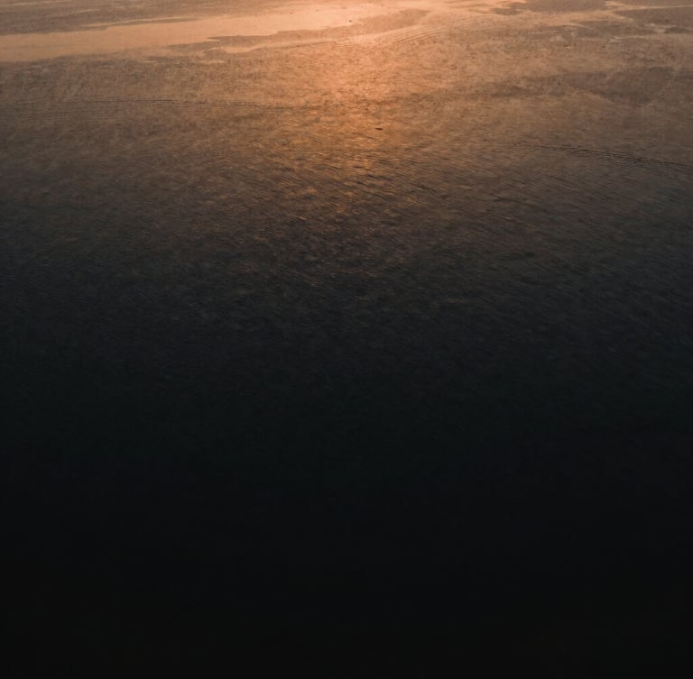 Cinematic wide angle drone view of Marano Lagunare lagoon, quiet dark water, golden hour light reflecting on the surface, deep shadows, rich ochre tones, minimalist composition, 35mm anamorphic lens