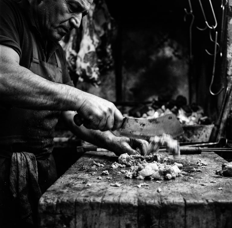 Ustanın zırh bıçağıyla eti kıyarken çekilmiş yüksek kontrastlı, dramatik fotoğrafı. Siyah-beyaz tonlarda, ustanın elleri ve bıçak ön planda, hareket bulanıklığıyla zanaatın enerjisi vurgulanmış. Düşük ışık, keskin detaylar, 50mm.
