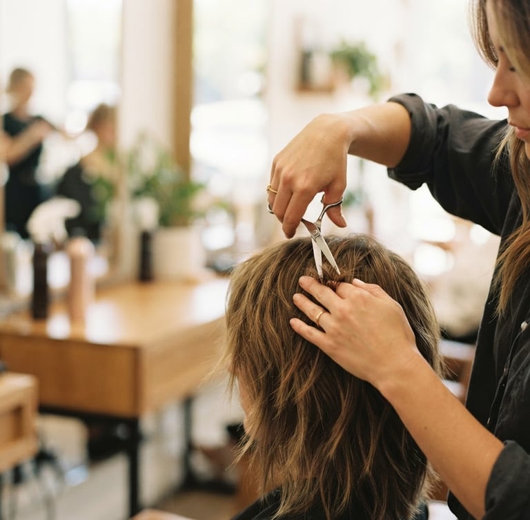 Close-up of a stylist's hands meticulously detailing hair with scissors, warm bright indoor light, modern salon setting, shallow depth of field, subject on the right third.