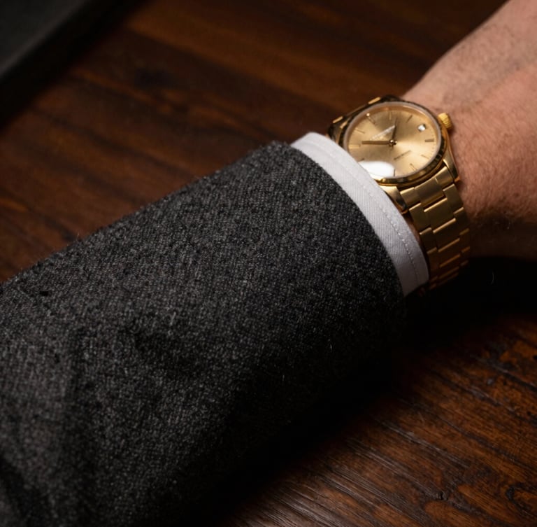 Tight macro detail of a tailored charcoal wool cuff and a gold watch against a dark wood table, low-key warm lighting, atmospheric