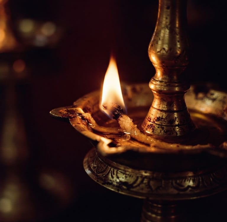 Macro close-up of a traditional Kerala brass oil lamp glowing with warm flickering light, deep maroon shadows, antique gold reflections, 35mm photography
