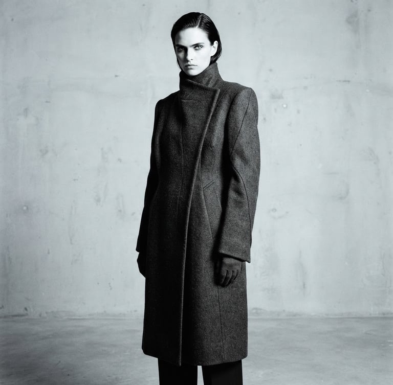 High-contrast minimalist fashion photography, model in structural charcoal wool coat, sharp directional studio lighting, desaturated cool tones, negative space