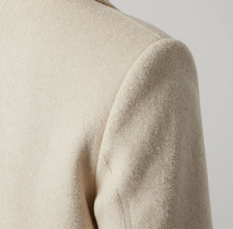 Extreme close-up of a tailored cream-colored wool coat shoulder seam, soft diffused studio lighting, high-contrast neutral tones, macro texture detail.