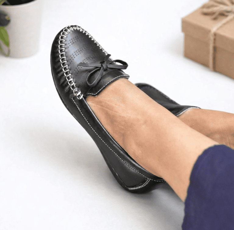 Mocasines cómodos de cuero negro para mujer con costuras blancas y un lazo decorativo.