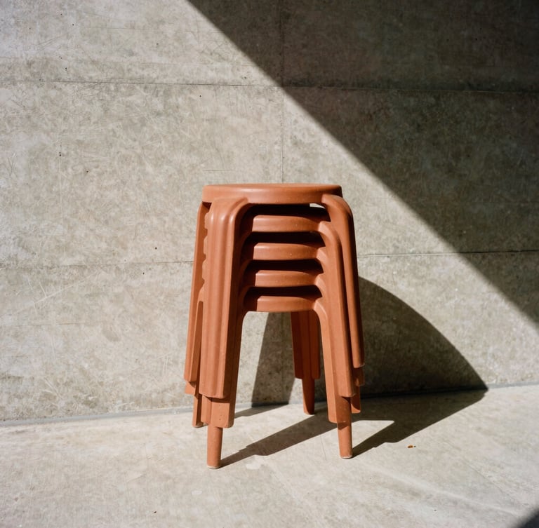 Low-angle architectural photograph of terracotta-toned stackable resin stools against a raw board-formed concrete wall, sharp geometric midday shadows, 35mm