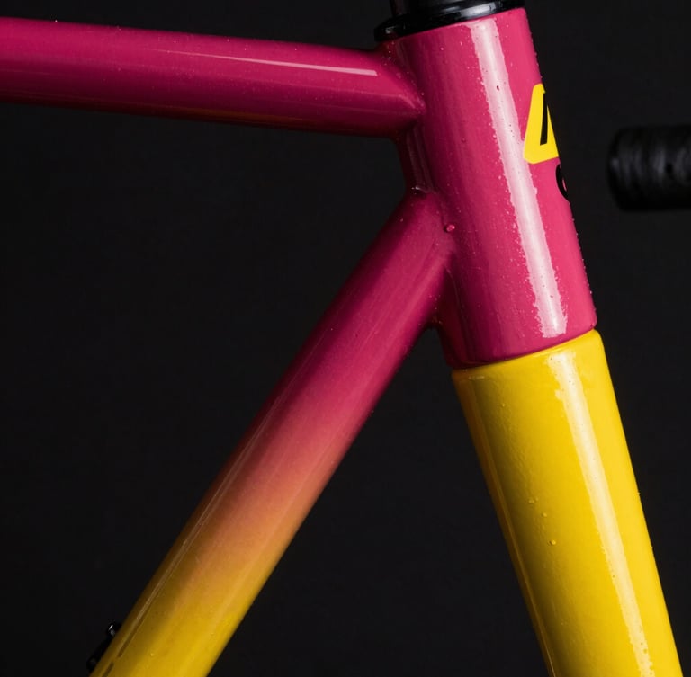 Extreme close-up of a bicycle frame headtube being painted with a deep magenta to electric yellow gradient, fine spray mist catching a sharp spotlight, dark background.
