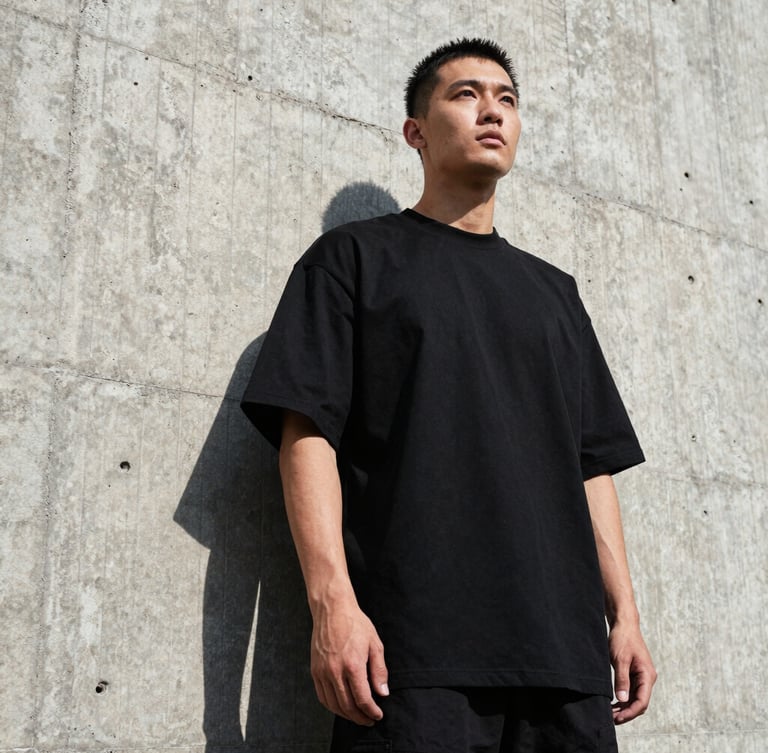 Low-angle heroic shot of a man wearing a heavy-weight black oversized t-shirt, standing against a raw concrete brutalist wall, harsh direct sunlight, high-contrast shadows