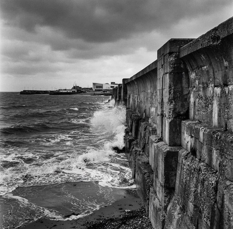 Wide-angle black and white film photograph of the brutalist Margate sea wall on an overcast day, high-contrast dark sea-navy water crashing, raw texture, negative space on the left
