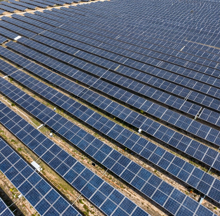 Wide aerial drone shot of massive utility-scale solar arrays stretching to the horizon under harsh midday sun, emphasizing scale and physical reality.