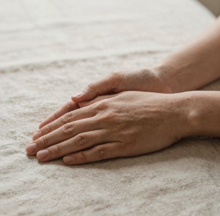 Close-up of hands gently resting on a textured linen surface, soft diffused morning light, shallow depth of field, conveying calm and grounded presence, warm editorial photography, earthy muted tones.