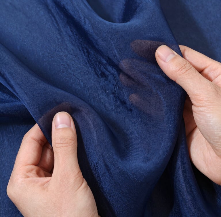 Macro overhead flat-lay of hands holding a premium Turkish organza swatch in deep navy blue, soft side-lit daylight highlighting the delicate weave and translucent texture