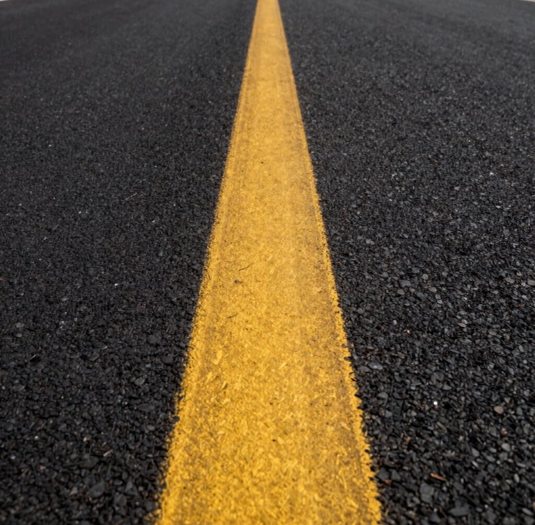 Low-angle perspective shot looking down a long, perfectly straight yellow traffic line on fresh black asphalt, bright morning light casting sharp textures.