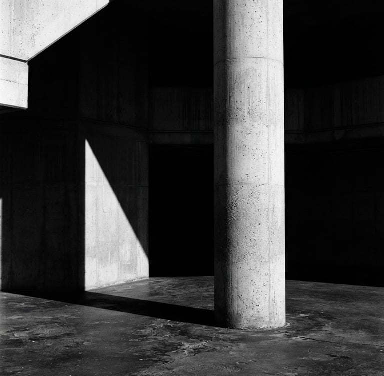 Stark black and white fine art photography, harsh high-contrast directional light, deep architectural shadows, concrete column silhouette, minimalist composition, 35mm film.