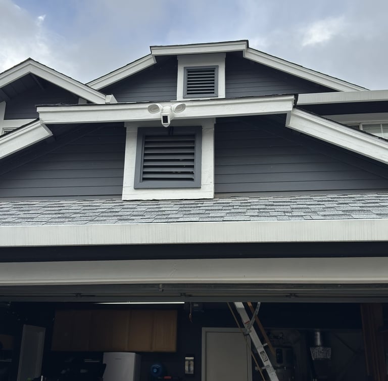 Outdoor home security camera and floodlight mounted on a gray house exterior above a garage.