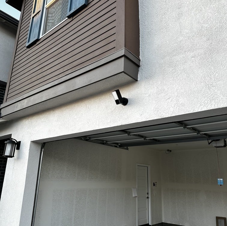 A black outdoor security camera mounted on a white stucco wall above a residential garage door.