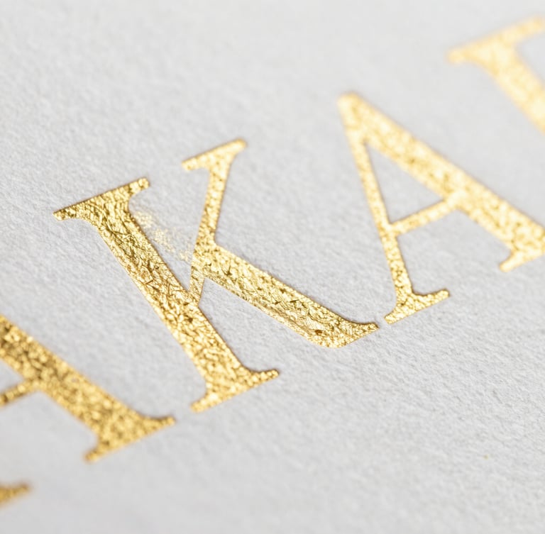 Macro close-up of heavy-weight textured paper with embossed gold foil lettering, crisp edges, bright clean studio lighting, shallow depth of field.