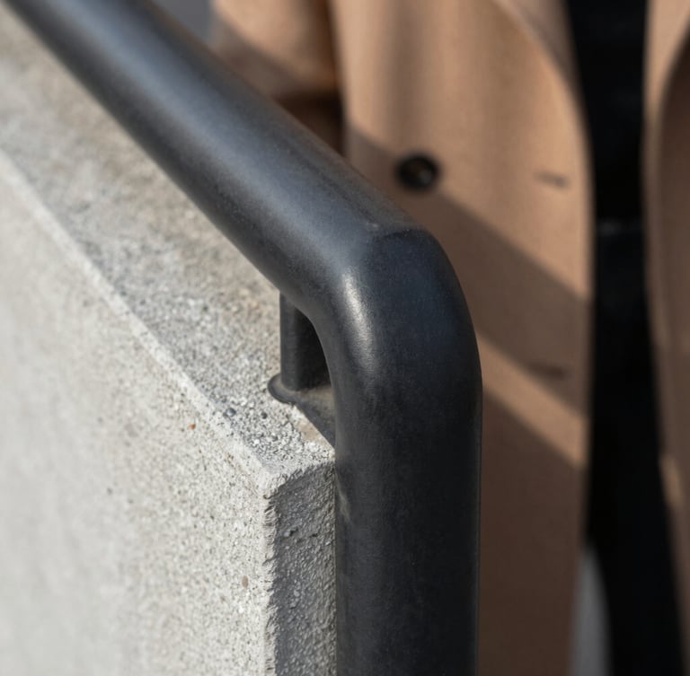Extreme close-up of a matte black steel handrail meeting raw concrete grain, soft natural light highlighting the texture, a coat draped casually in the background.