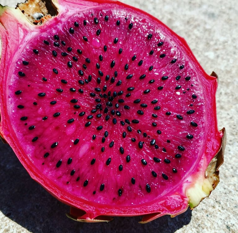 Macro close-up of a sliced red dragon fruit showing vibrant pink flesh and tiny black seeds, glistening with moisture on a neutral stone surface under high-contrast sunlight.