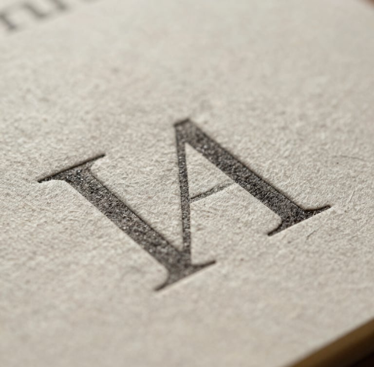 Extreme macro crop of heavy cotton paper stock with deep debossed typography under soft, diffused north daylight, showing rich tactile texture.