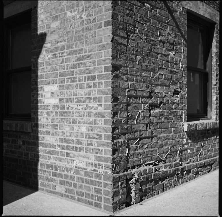 High-contrast monochrome medium-format photograph of a weathered brick corner in a shifting urban neighborhood, harsh midday shadows, visible film grain, unposed.