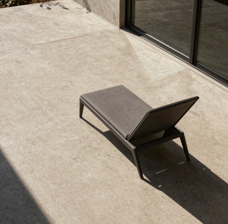 Wide architectural environmental shot, hard direct sunlight casting dramatic shadows on a raw concrete deck, a single charcoal structural lounge chair positioned near a glass threshold, medium format.