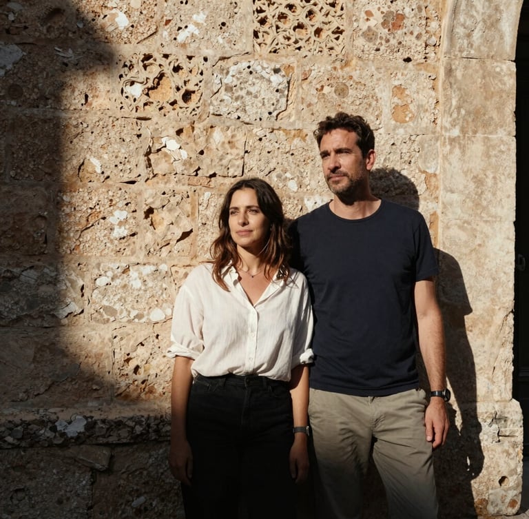 Cinematic medium-format portrait of a couple standing by an ancient stone wall in Mallorca, harsh midday sun casting deep shadows, warm Mediterranean light, raw candid emotion, look away from camera.