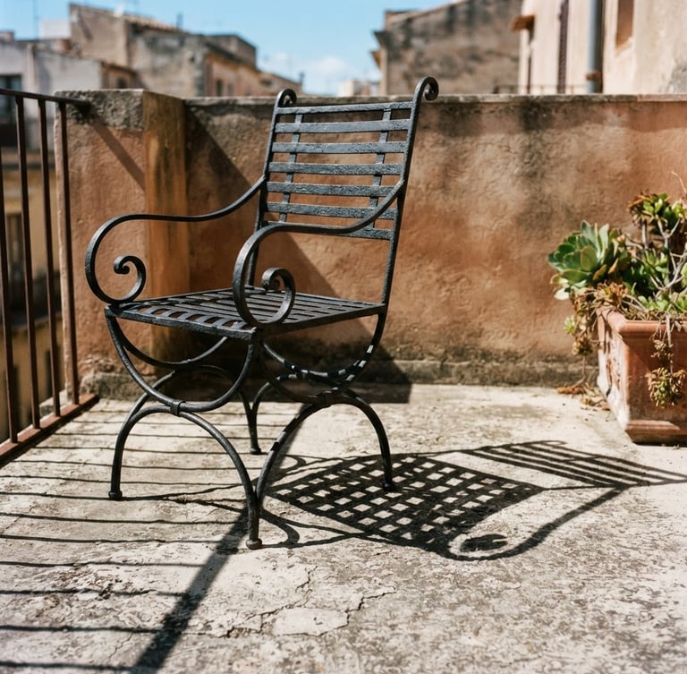 Architectural editorial crop of a heavy charcoal iron balcony chair, casting sharp geometric shadows on weathered concrete, warm terracotta accent tones, 35mm photography