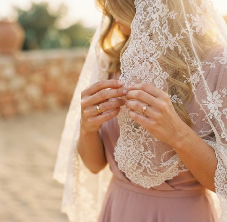 A close-up of a bride's hands adjusting a delicate lace veil, warm golden hour light filtering through, soft dusty rose tones, elegant editorial style.