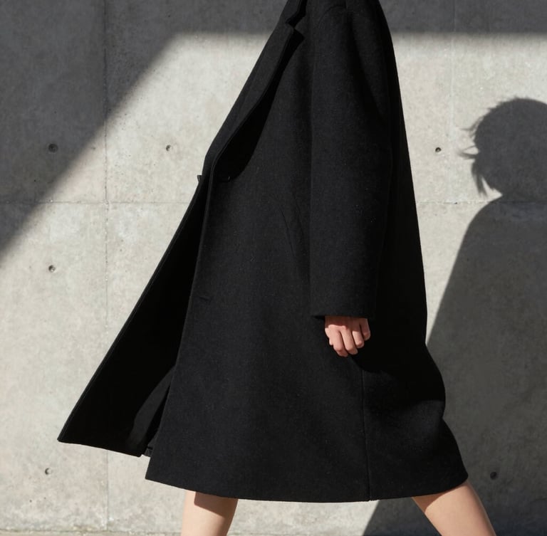 High-contrast architectural crop of a model in motion, wearing an oversized structural black coat, harsh direct sunlight, casting sharp geometric shadows on a concrete wall.