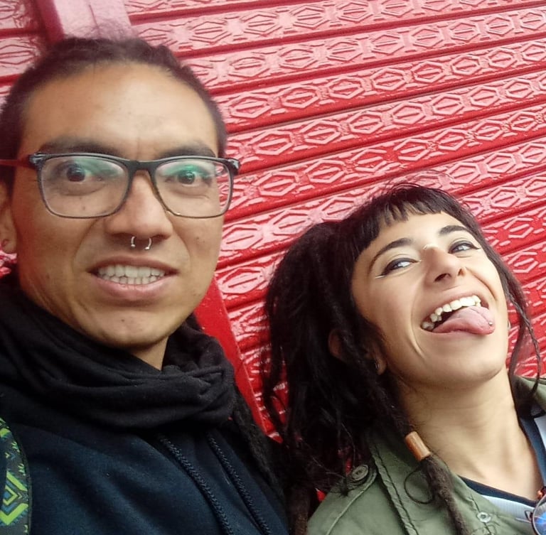 Un hombre sonriente con gafas y una mujer sacando la lengua, posando frente a una pared roja con tex
