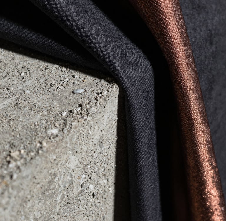Macro photograph of rough-hewn raw concrete meeting heavy, dark, hand-stitched textile fabrics, high-contrast side lighting with deep shadows and metallic copper highlights.