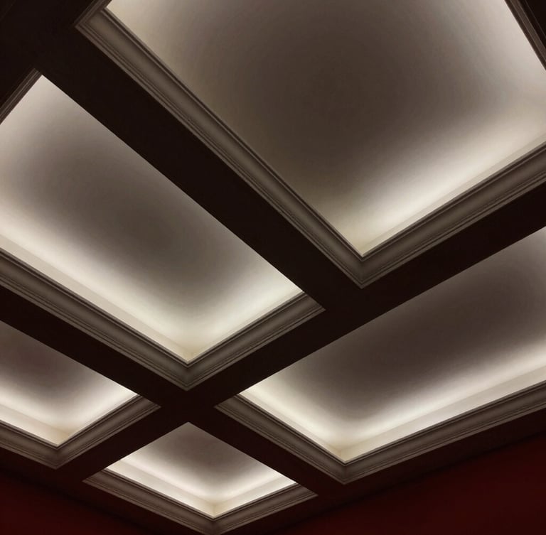 Low-angle architectural photograph looking up at a pristine, geometric white coffered ceiling with integrated warm lighting, dramatic raking light highlighting crisp millwork joints, high-contrast charcoal shadows, 35mm lens.