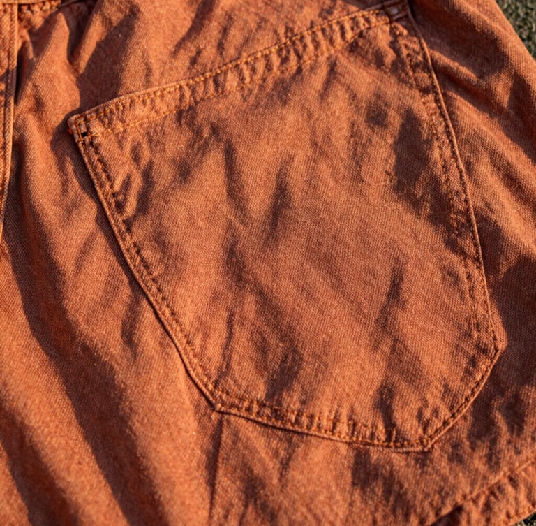 Close-up of worn terracotta-toned denim fabric showing beautiful textured fade lines and sturdy double-stitching in warm golden-hour sunlight.
