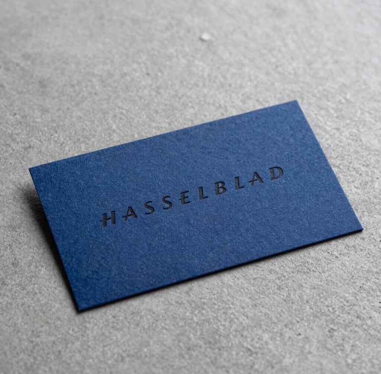 Macro close-up of premium heavy-weight cotton business cards with deep debossed black typography on a cool grey concrete surface, sharp studio lighting, minimal composition