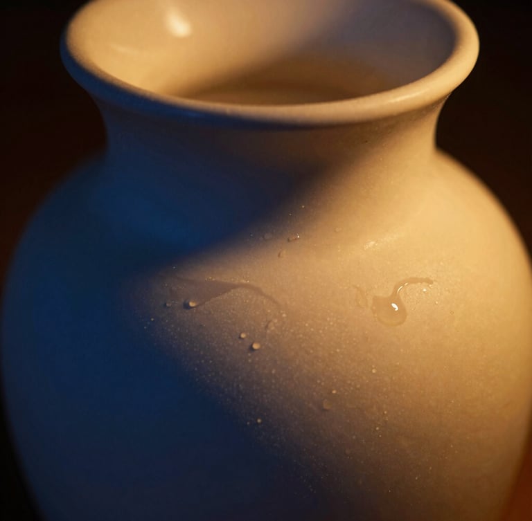 Low-key warm golden candlelight casting deep blue shadows over an elegant ceramic vessel, macro texture of melted wax, shallow depth of field.