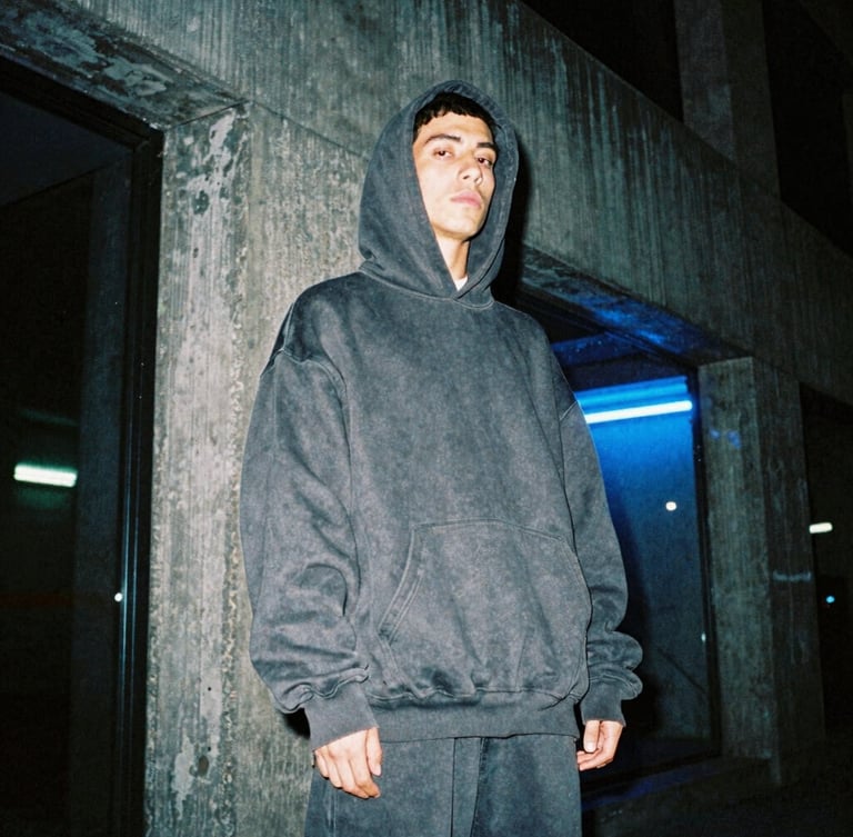 Moody low-angle shot of a hip-hop artist in an oversized heavy-weight dark grey hoodie standing in a brutalist concrete alley in Mexico City at night, midnight blue neon reflections, 35mm film.