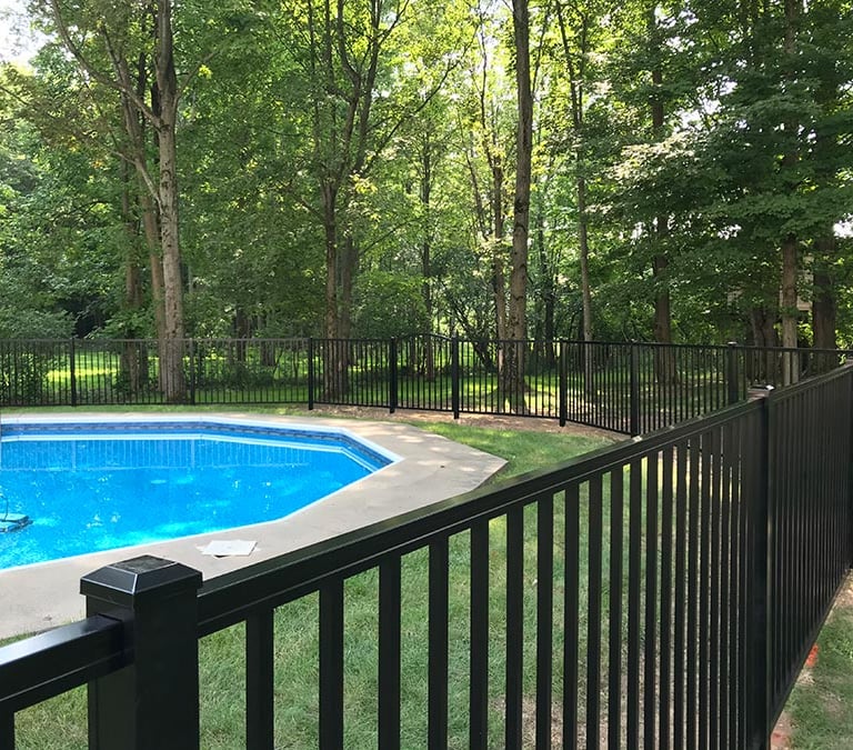 Black aluminum safety fence surrounding an inground swimming pool in a lush wooded backyard.