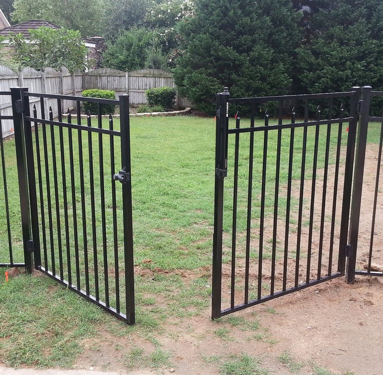 Open black ornamental aluminum fence gate leading to a green grassy backyard with a wooden privacy fence.