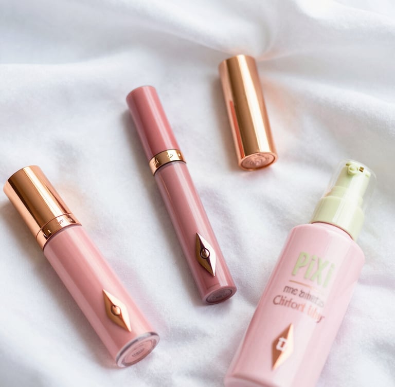 Overhead flatlay of premium pink cosmetics, Charlotte Tilbury and Pixi bottles on soft white silk, diffused natural morning light, shallow depth of field