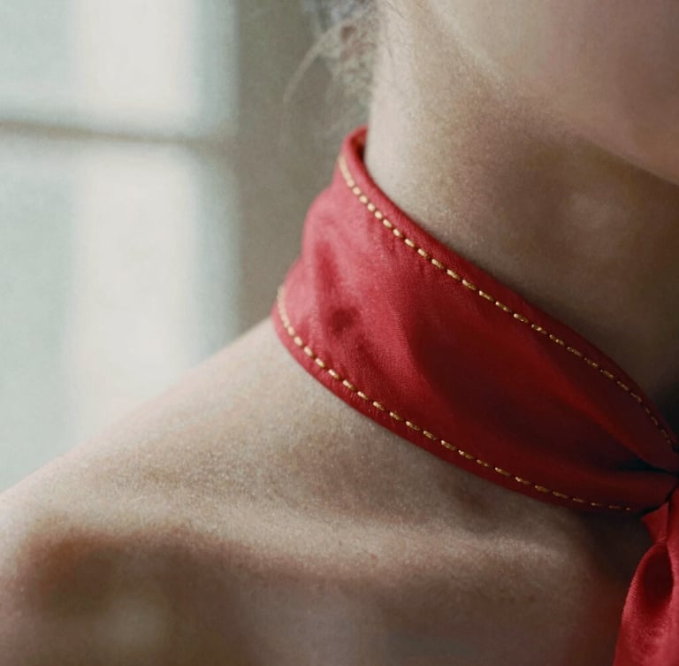 Extreme close-up of delicate gold hand-stitching on a scarlet silk collar, soft directional natural light with deep soft shadows, 35mm film grain
