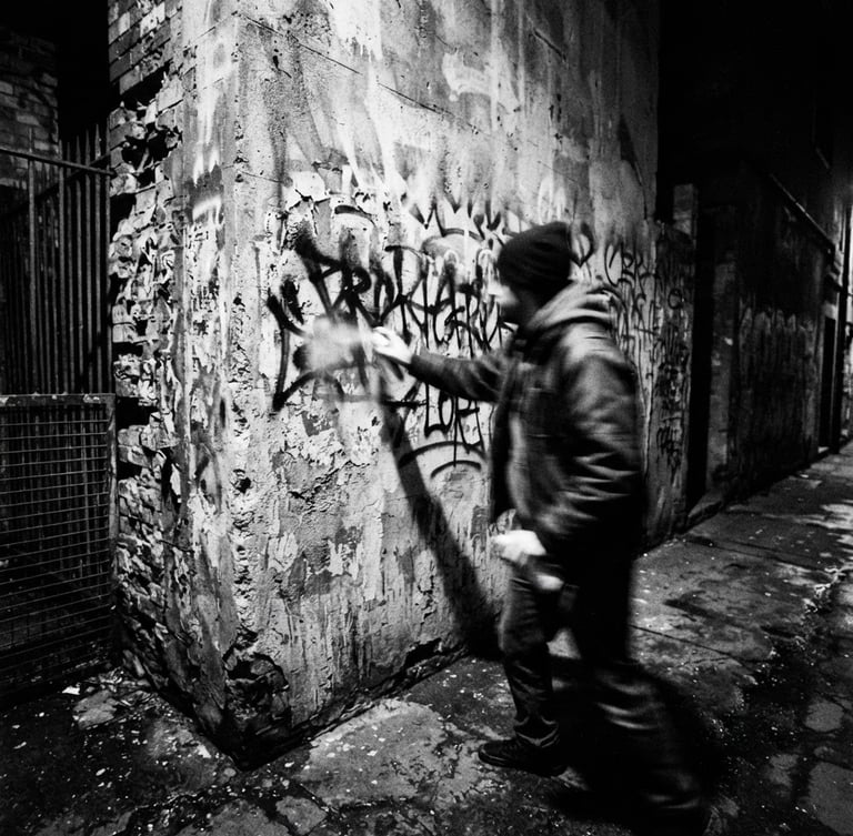 Raw flash photography, high contrast black and white, gritty urban environment, direct harsh light, 28mm wide-angle lens, showing a blurred figure spray-painting a defiant message on a concrete wall.