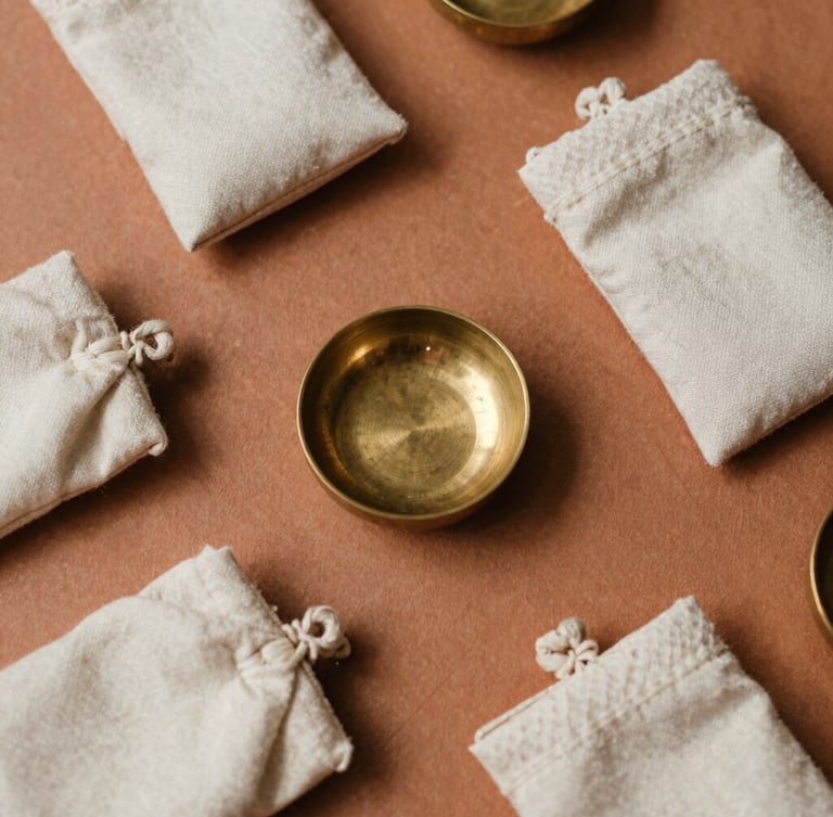 Overhead flat-lay of hand-woven raw silk pouches and small, heavy-milled brass incense holders on a warm clay surface, soft diffused lighting.