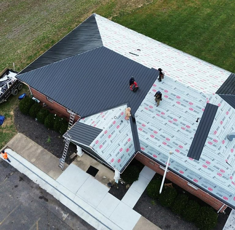 Aerial view of contractors installing a new black metal roof on a residential home with protective underlayment.