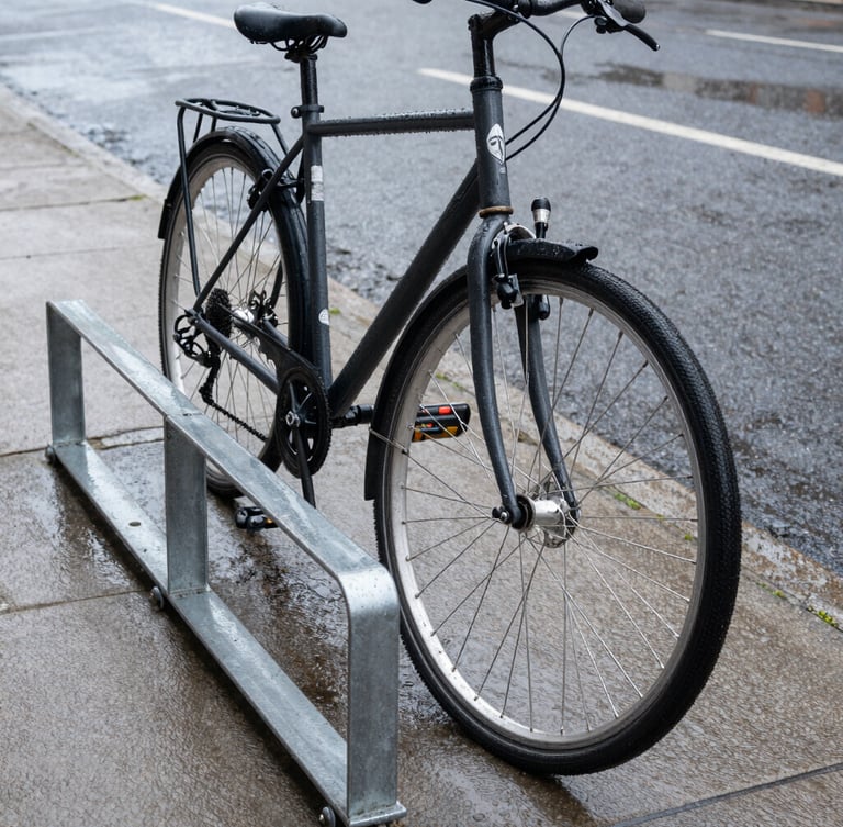 Low-angle street perspective of a utility commuter bike locked to a heavy steel rack on a wet asphalt sidewalk, overcast daylight, water droplets on the frame.