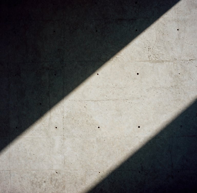 A 35mm film frame of a concrete wall cast in harsh diagonal shadow, high-contrast natural light, desaturated tones