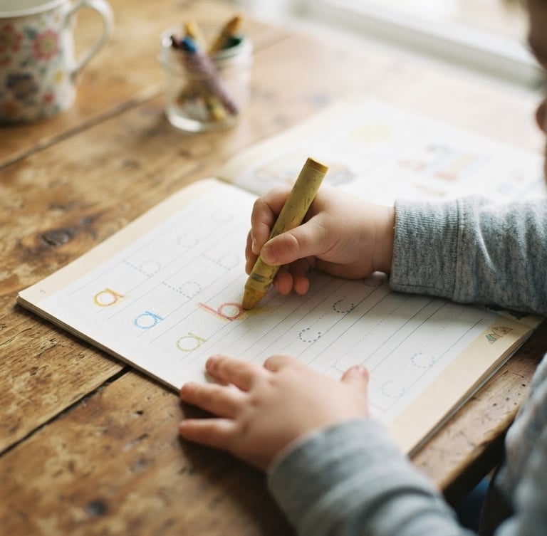 Overhead shot of children's hands holding a thick crayon, tracing letters on a printed workbook page, on a warm wooden kitchen table, soft diffused morning daylight, gentle muted pastel tones, shallow depth of field, 35mm film look