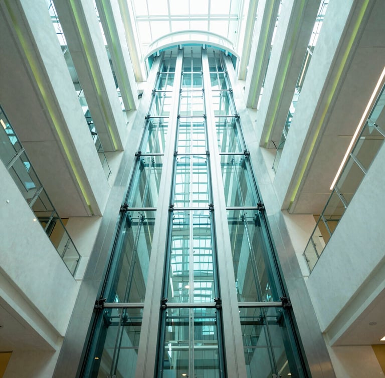 Low-angle wide shot looking up a sleek glass elevator shaft rising through a sunlit corporate atrium, cool teal reflections, bright high-key lighting, architectural symmetry.